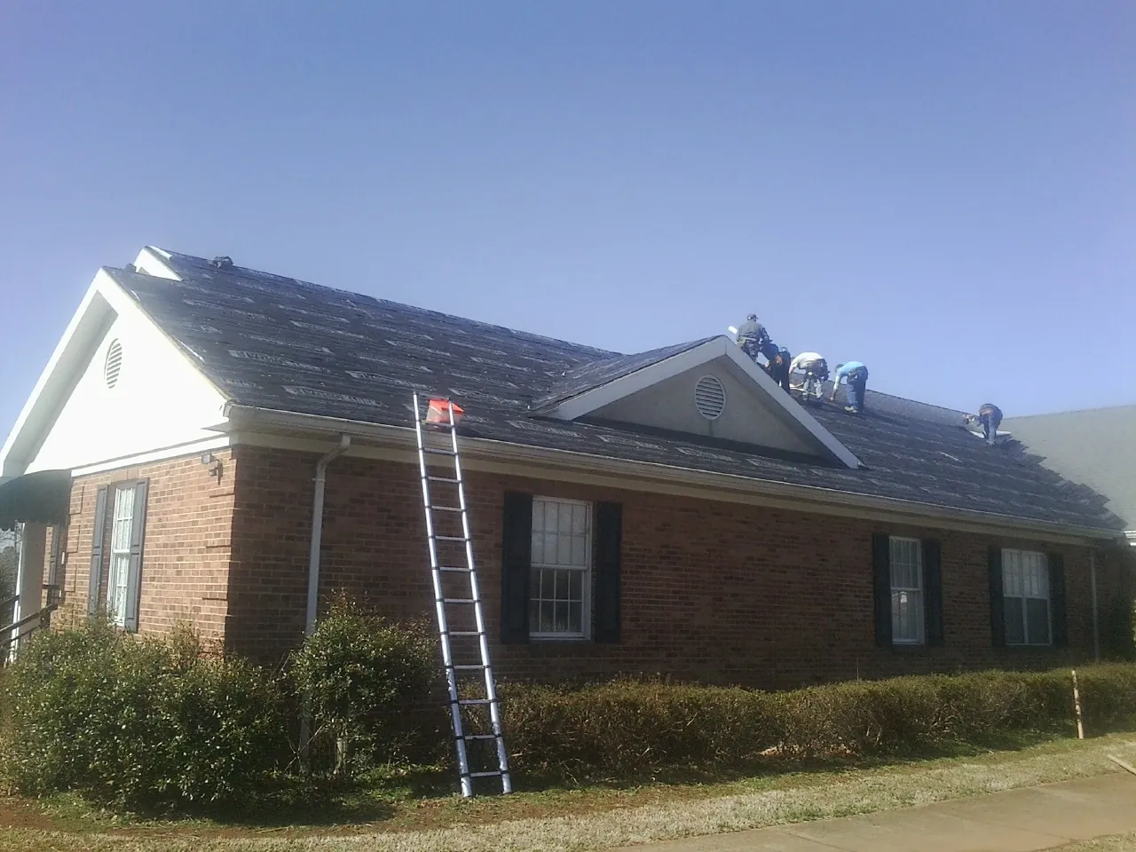 Crew tearing shingles off a brick ranch roof with a ladder set — Emergency Roof Repair work by our crew in Vass