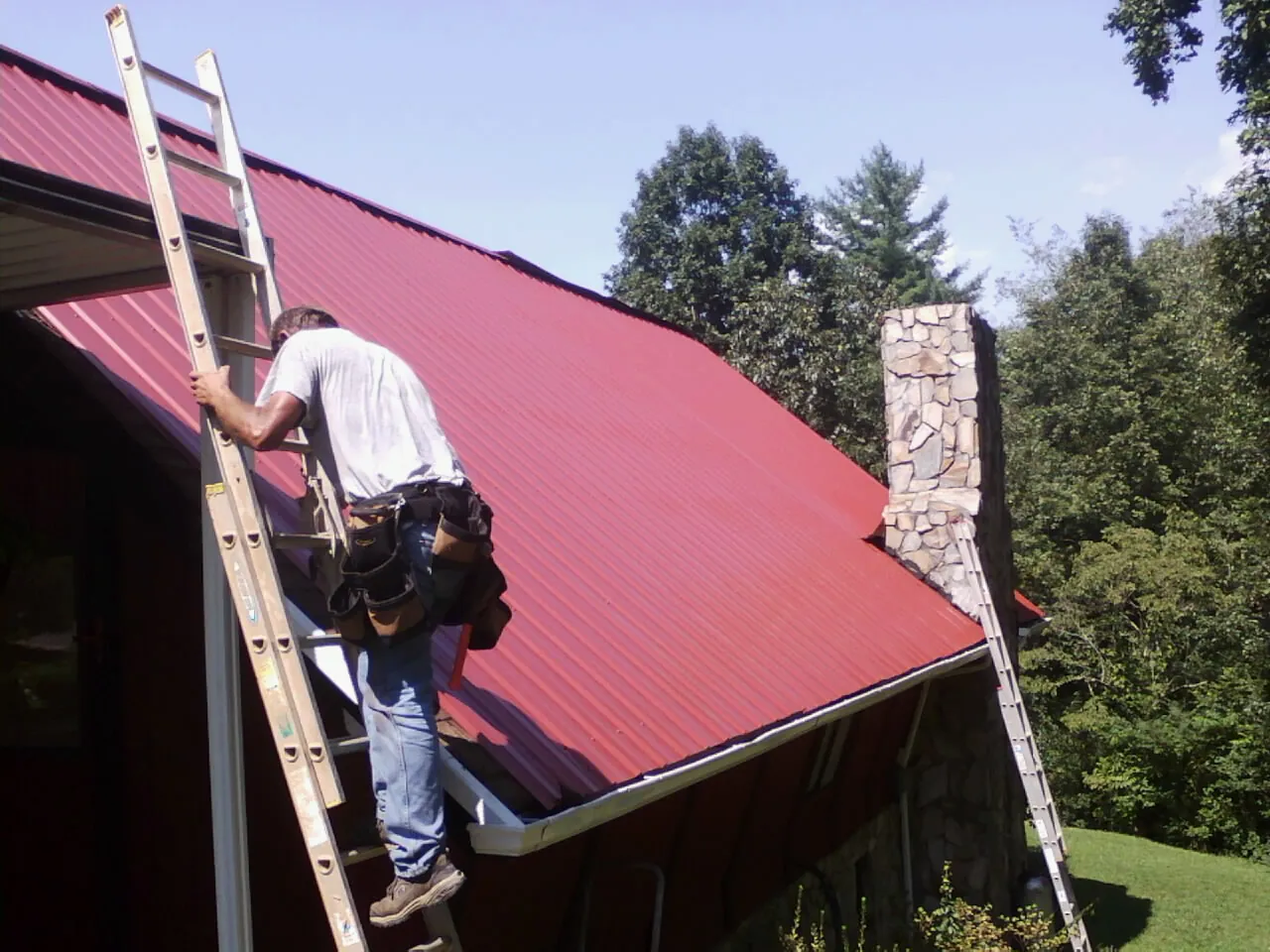 Roofer on a ladder beside a red metal roof and stone chimney — Soffit & Fascia Repair by Roofing Brothers in Vass